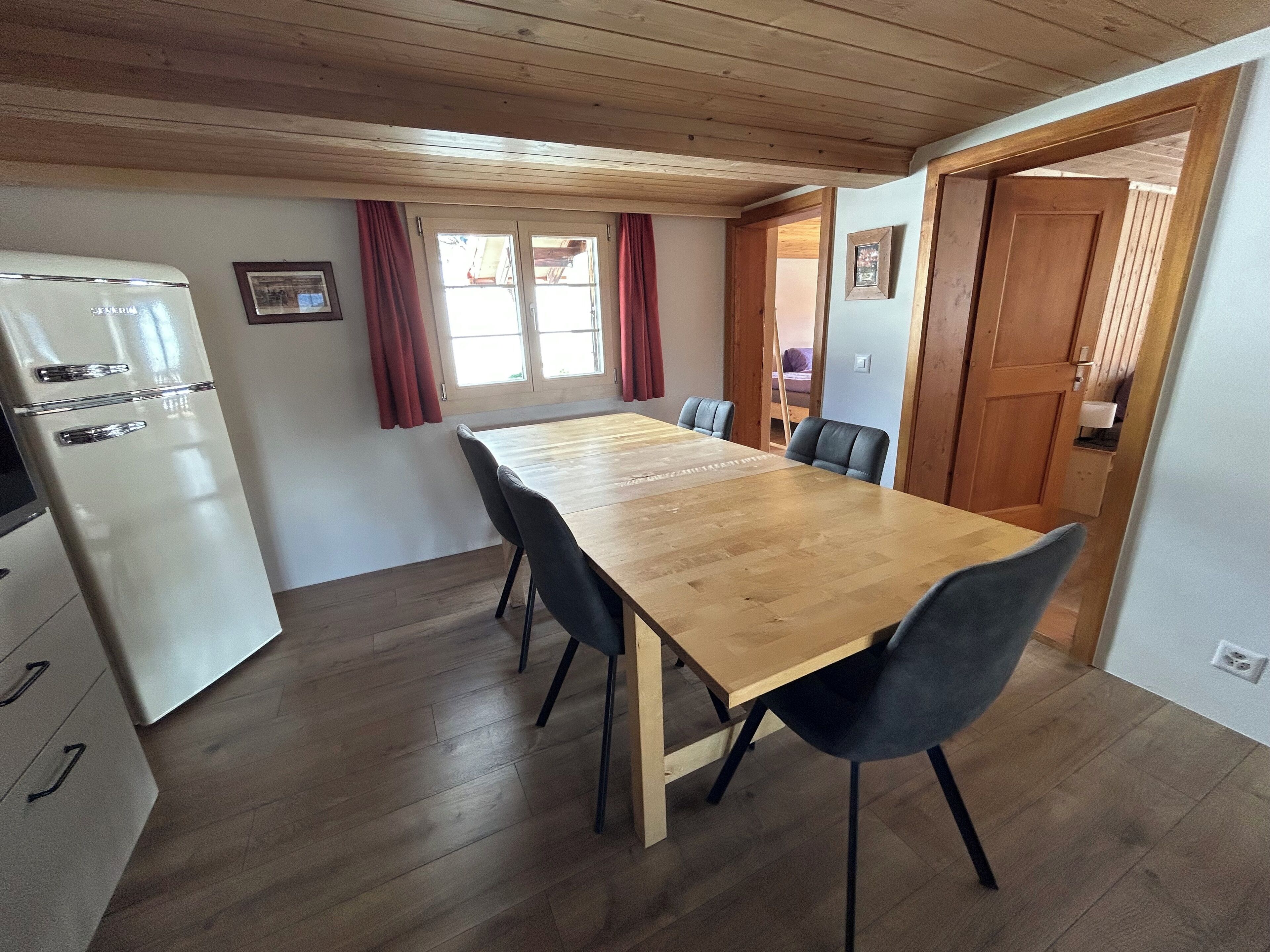 Een kleine keuken met een houten tafel en stoelen, een witte koelkast, en een raam met gordijnen.
