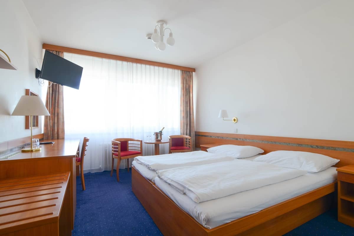 A hotel room with two beds, a desk with a chair, a TV mounted on the wall, and a window with curtains.