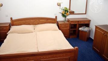 Un lit en bois avec une tête de lit, une table de chevet avec une lampe, un miroir, une petite armoire en bois et un sol recouvert d’une moquette bleue.