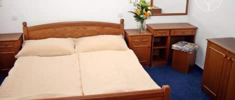 A wooden bed with a headboard, a bedside table with a lamp, a mirror, a small wooden cabinet, and a blue carpeted floor.