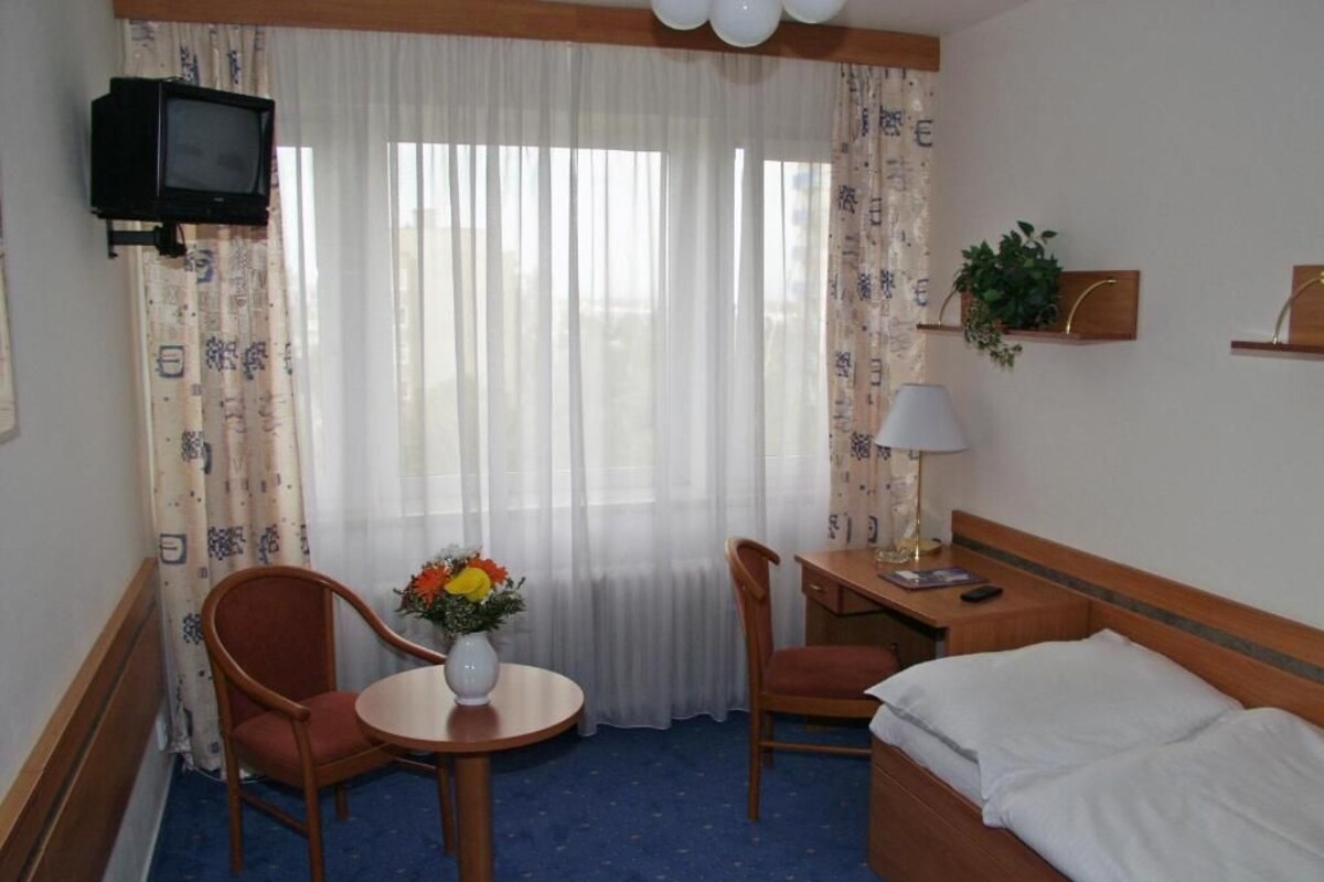 Ett hotellrum med en säng, två stolar, ett litet bord med en vas med blommor, ett skrivbord med en lampa, en tv och ett fönster med gardiner.
