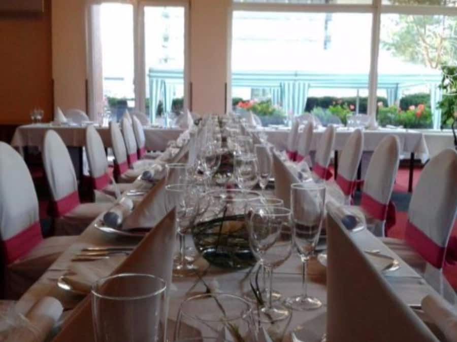 A long dining table set with chairs, glasses, and cutlery, in a room with large windows and a view of greenery outside.