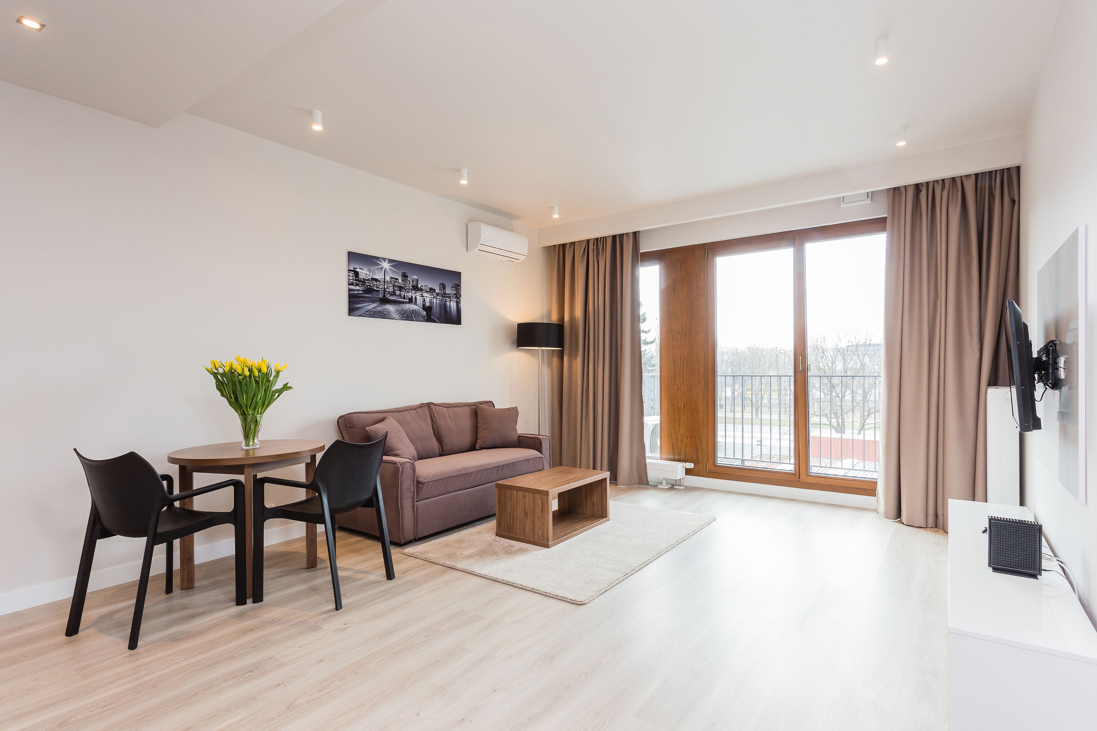 En moderne stue med sofa, sofabord og stole, et spisebord med en vase blomster, og et stort vindue med gardiner.