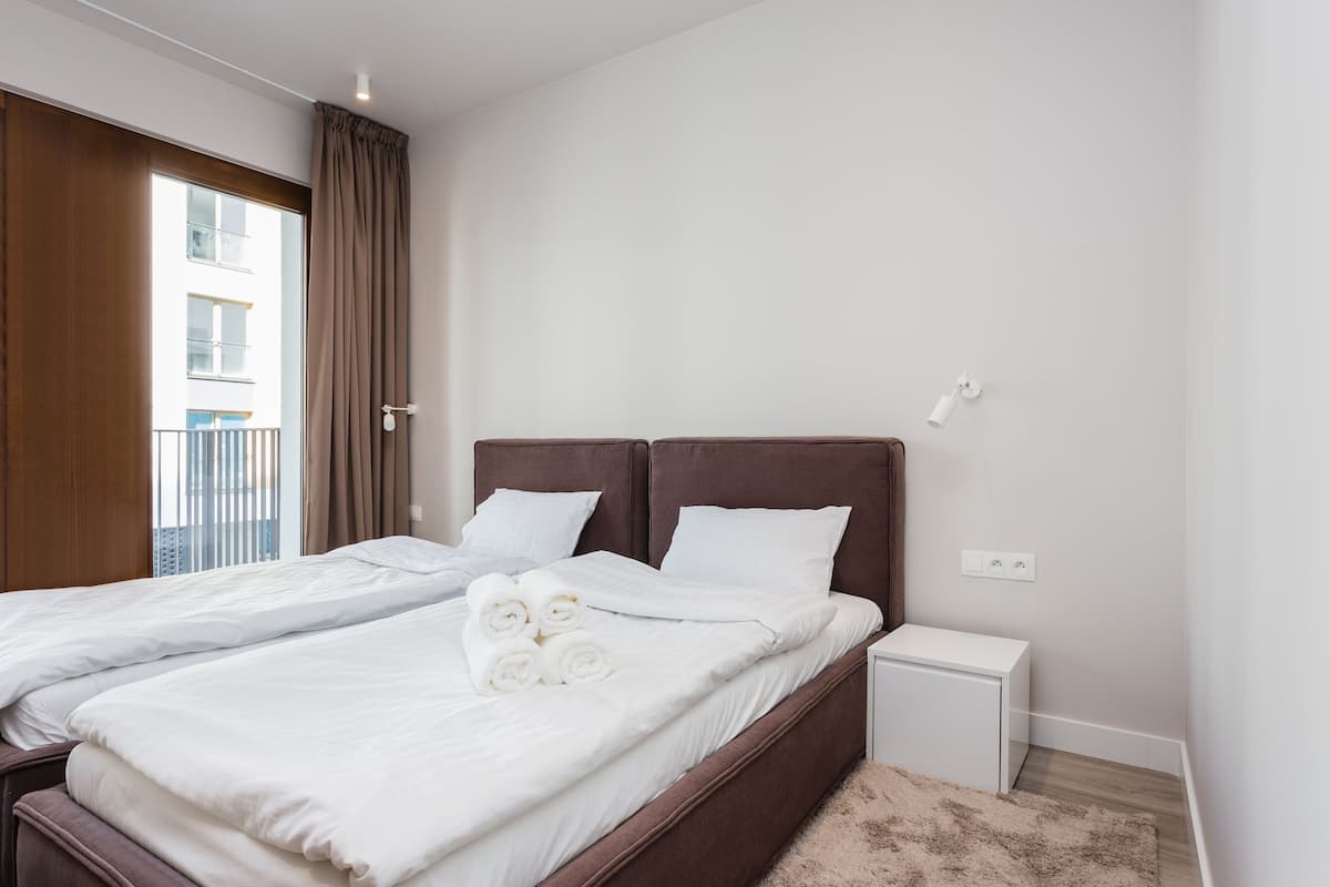 A double bed with white linens and two pillows, a bedside table, and a view of a building through a window with curtains.
