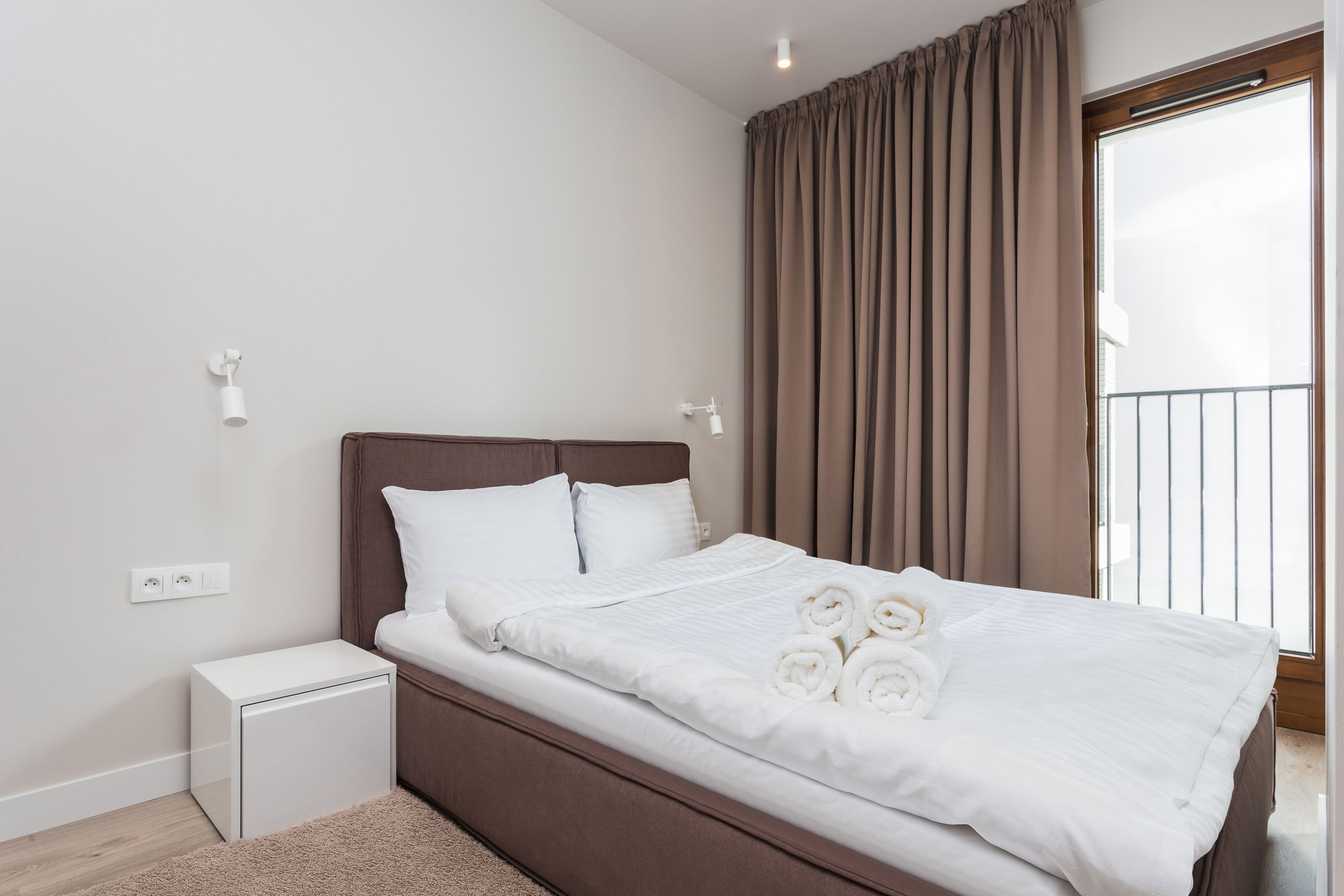 A neatly made bed with white linens and rolled towels, a small bedside table, and a power outlet on the wall.