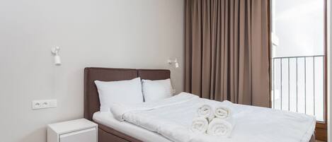 A neatly made bed with white linens and rolled towels, a small bedside table, and a power outlet on the wall.