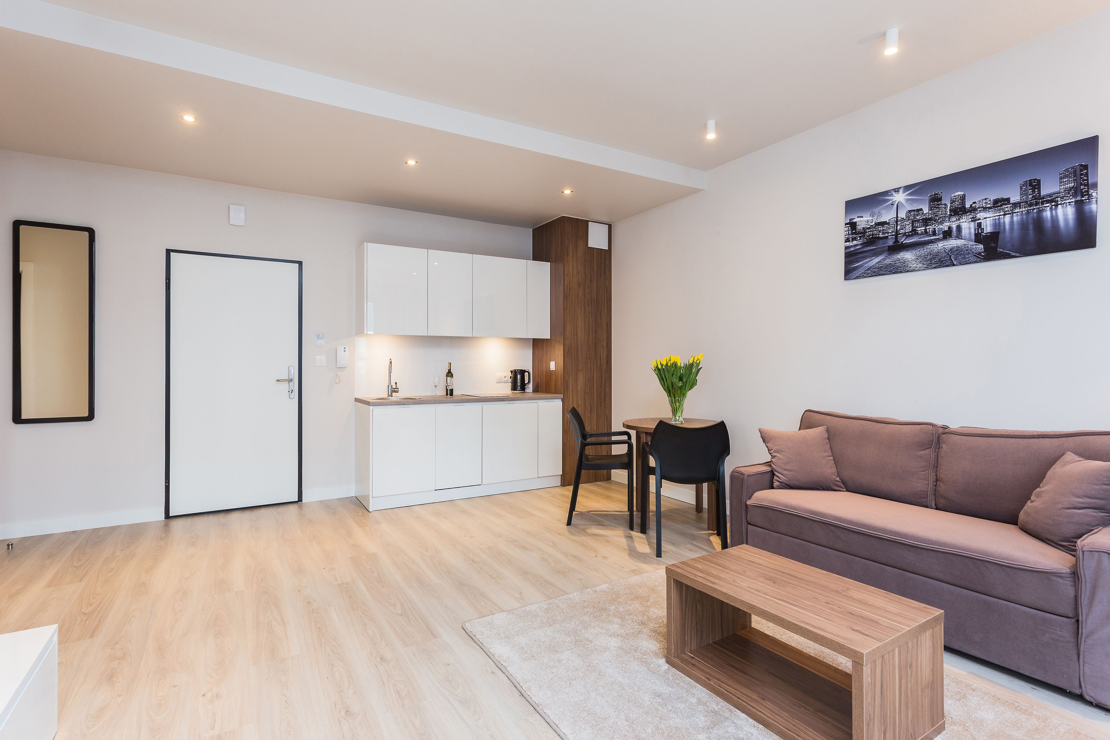 A modern apartment with a kitchenette, a living area with a sofa and coffee table, and a cityscape painting on the wall.