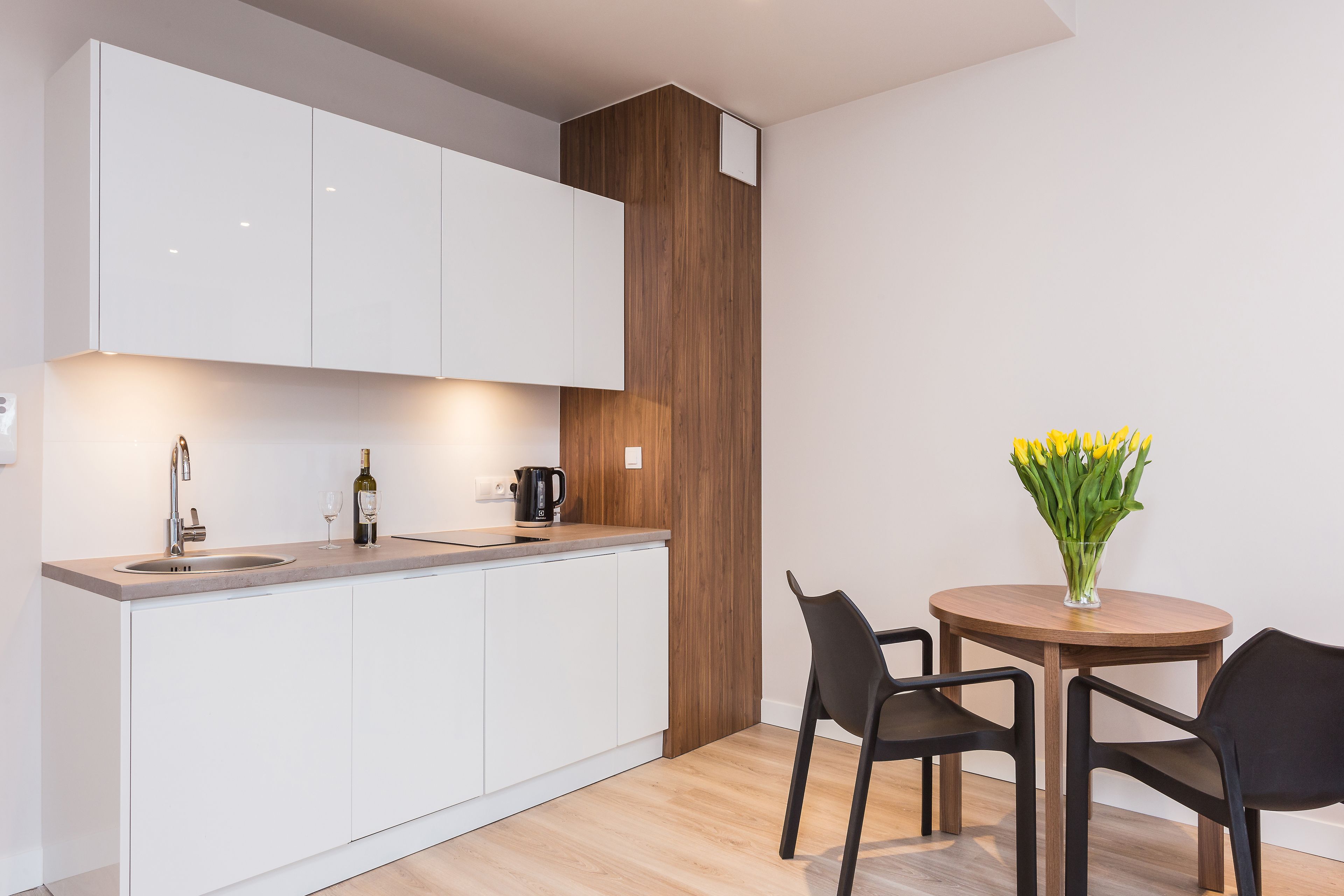 A modern kitchen with white cabinets, a wooden accent wall, a sink, and a coffee maker. There is a round wooden table with two chairs and a vase of yellow flowers.
