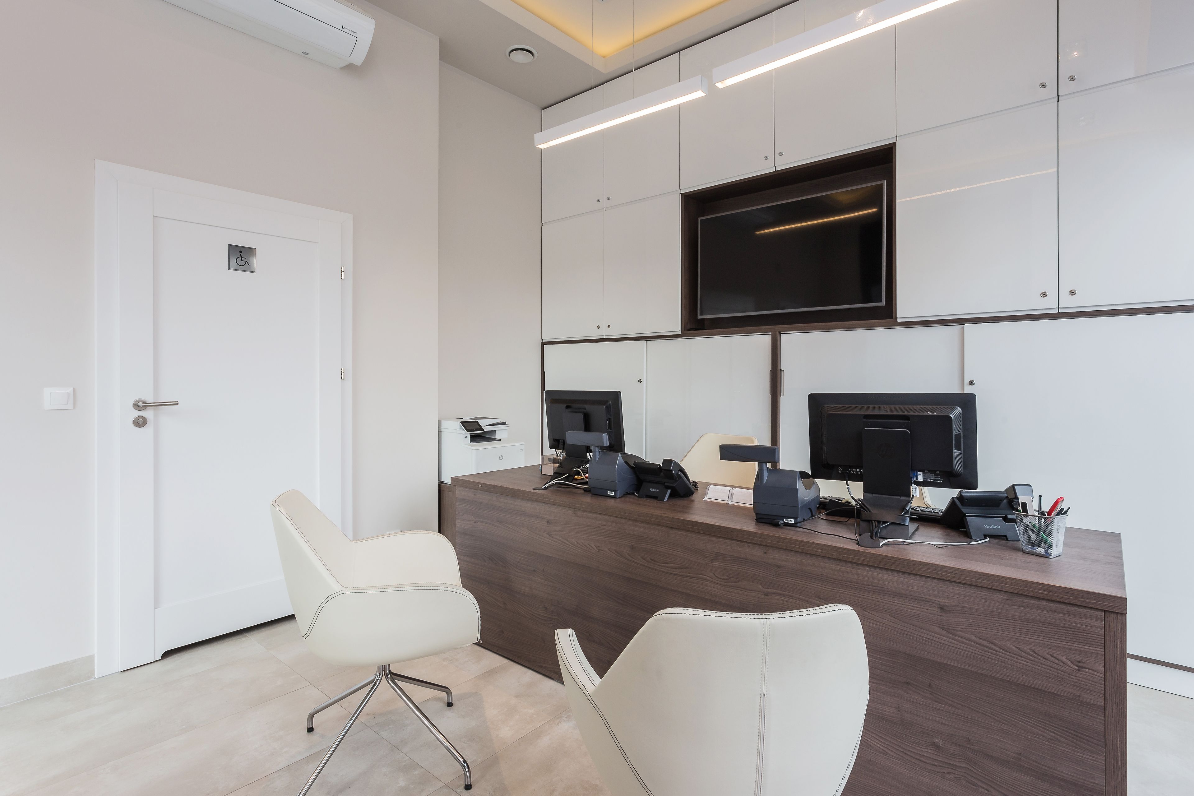A modern office space with a reception desk, two white chairs, and a large flat-screen TV mounted on the wall.