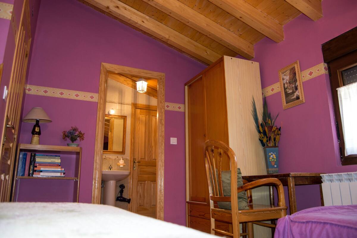 Una habitación con paredes moradas, vigas de madera en el techo, una cama, una silla de madera, una estantería con libros, una mesita con una lámpara y una ventana con cortina.
