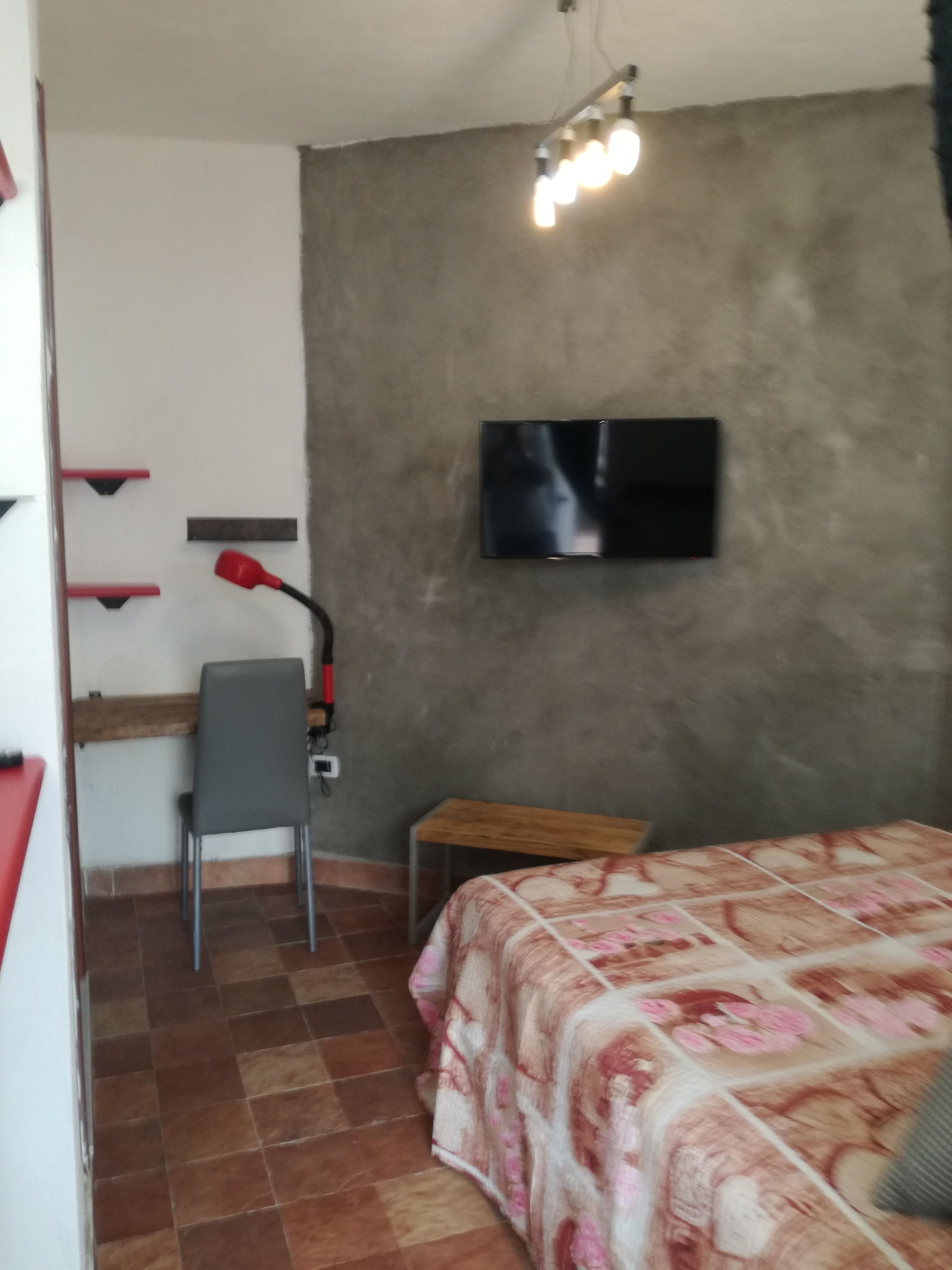 Un dormitorio con una cama, un escritorio, una silla, un televisor montado en la pared y una lámpara roja.