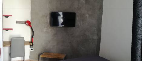 Quarto pequeno e compacto, com uma cama, uma escrivaninha e uma televisão fixada na parede.