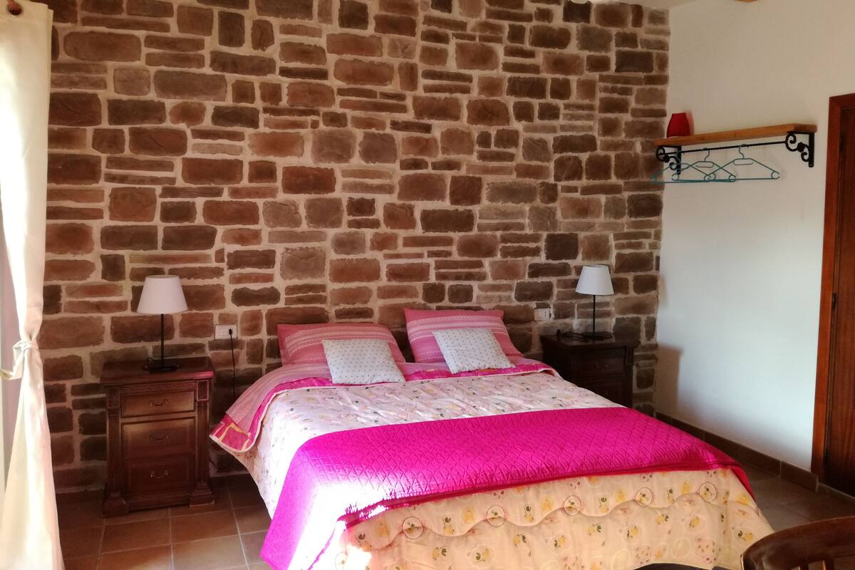 A bedroom with a stone wall, a bed with pink bedding, two bedside tables with lamps, and a wooden ceiling beam.