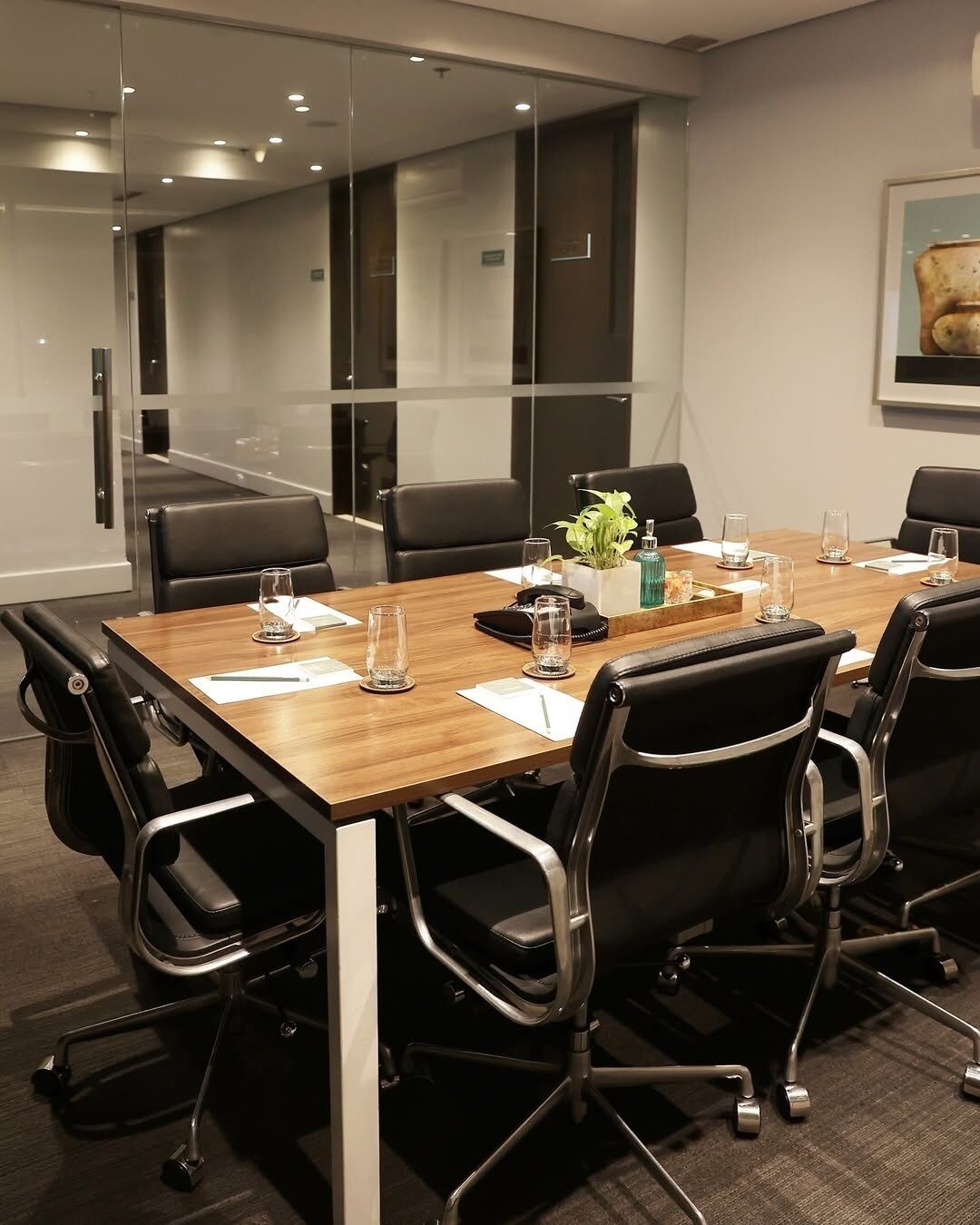 A conference room with a long table, black chairs, glass partitions, and a framed picture on the wall.