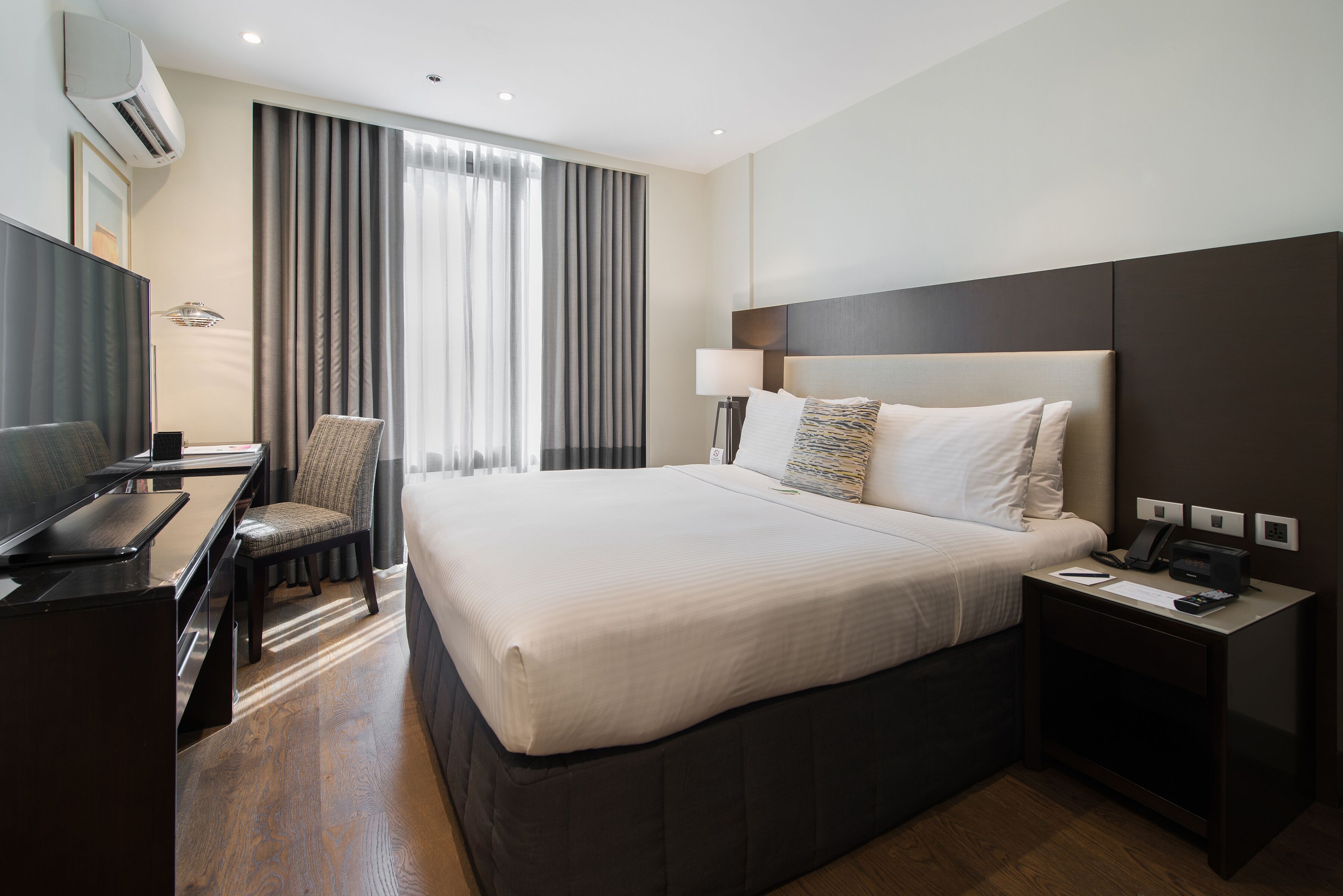 A hotel room with a large bed, a desk, a chair, a TV, and a bedside table with a lamp and phone.