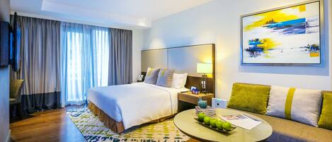 Habitación de hotel con una cama grande, un sofá, una mesa de centro con manzanas, un cuadro en la pared y un televisor a la izquierda.