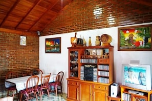 Un comedor con pared de ladrillo, techo de madera y una mesa con sillas. Hay cuadros en la pared y un mueble de madera con objetos encima.