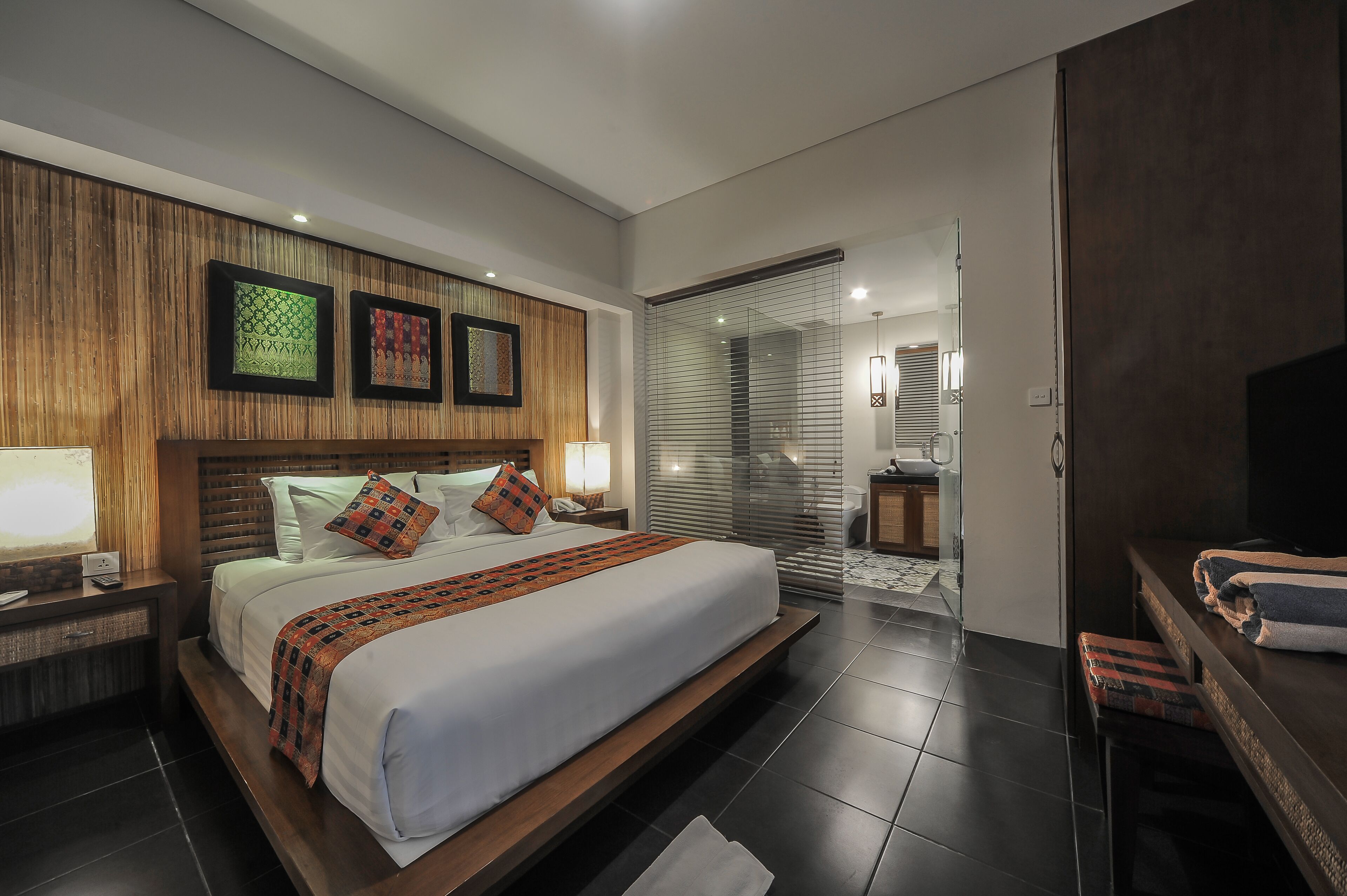 Une chambre d’hôtel moderne avec un grand lit, une tête de lit en bois, une table de chevet, une lampe, une salle de bain avec un lavabo et des toilettes, et un téléviseur.