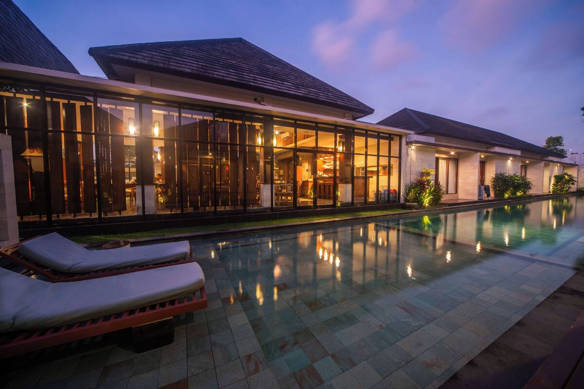 A modern hotel with a swimming pool, lounge chairs, and a well-lit interior.