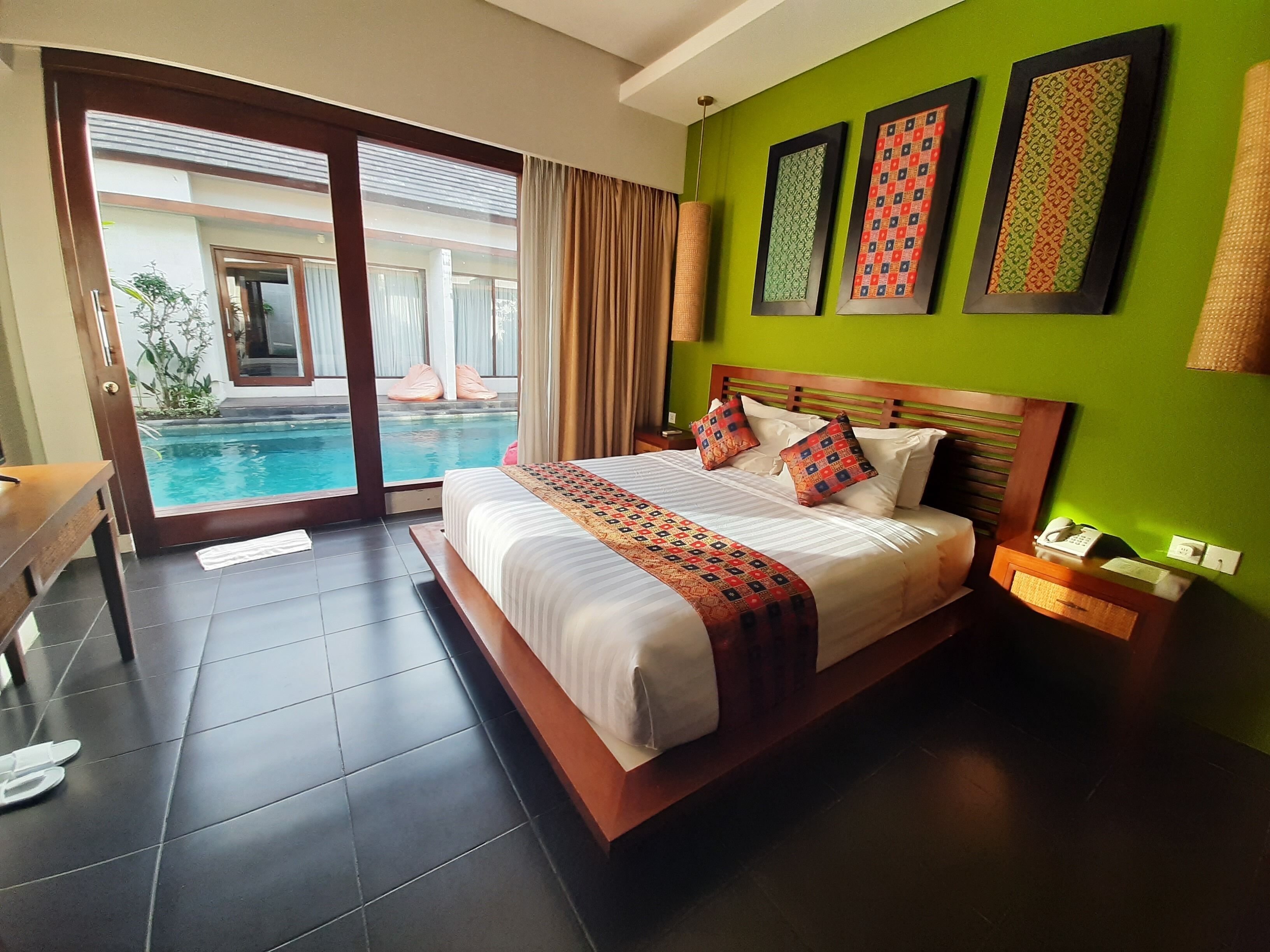 A hotel room with a large bed, a bedside table with a phone and a lamp, a green wall with framed artwork, and a view of a swimming pool through a glass door.