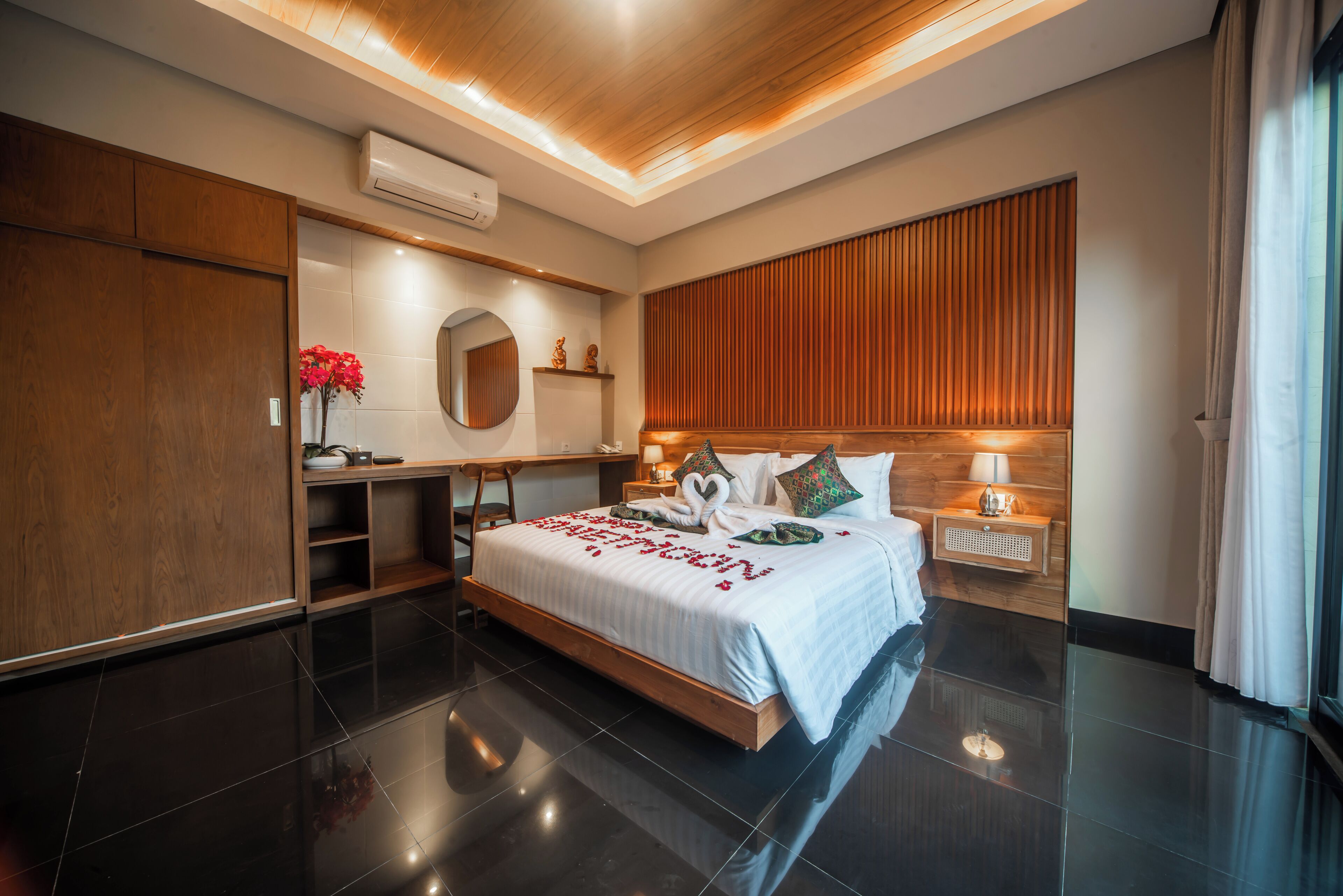 A modern hotel room with a large bed, wooden headboard, a bedside table with a lamp, a mirror, and a small shelf with decorative items.
