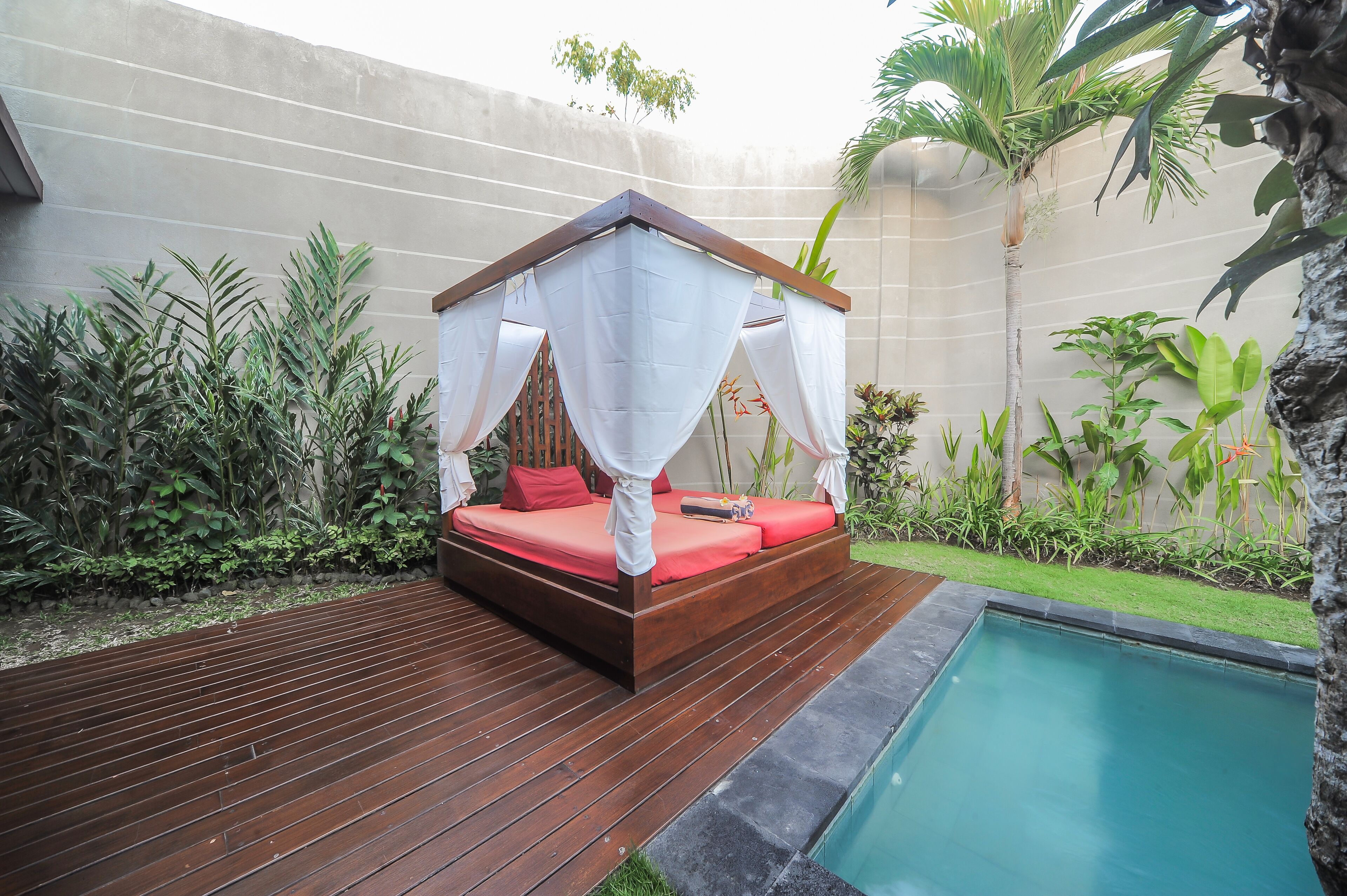 A wooden deck with a bed featuring a canopy, surrounded by lush greenery and a pool.