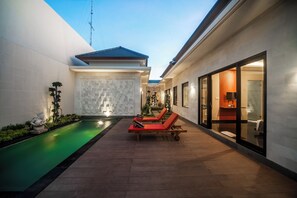 A modern outdoor pool area with lounge chairs, a small water feature, and a well-lit interior space with a large window. - Gaing Mas Jimbaran Villas by Gaing Mas Group (Jimbaran)