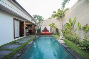 Una piscina con una estatua, un banco rojo y un pequeño jardín con plantas tropicales.