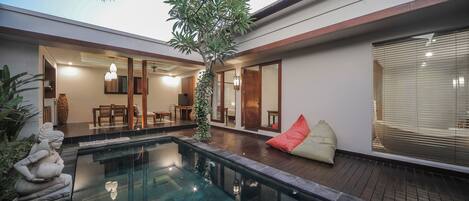 Un área de piscina exterior moderna con barandilla de vidrio, terraza de madera y zona de estar con cojines.