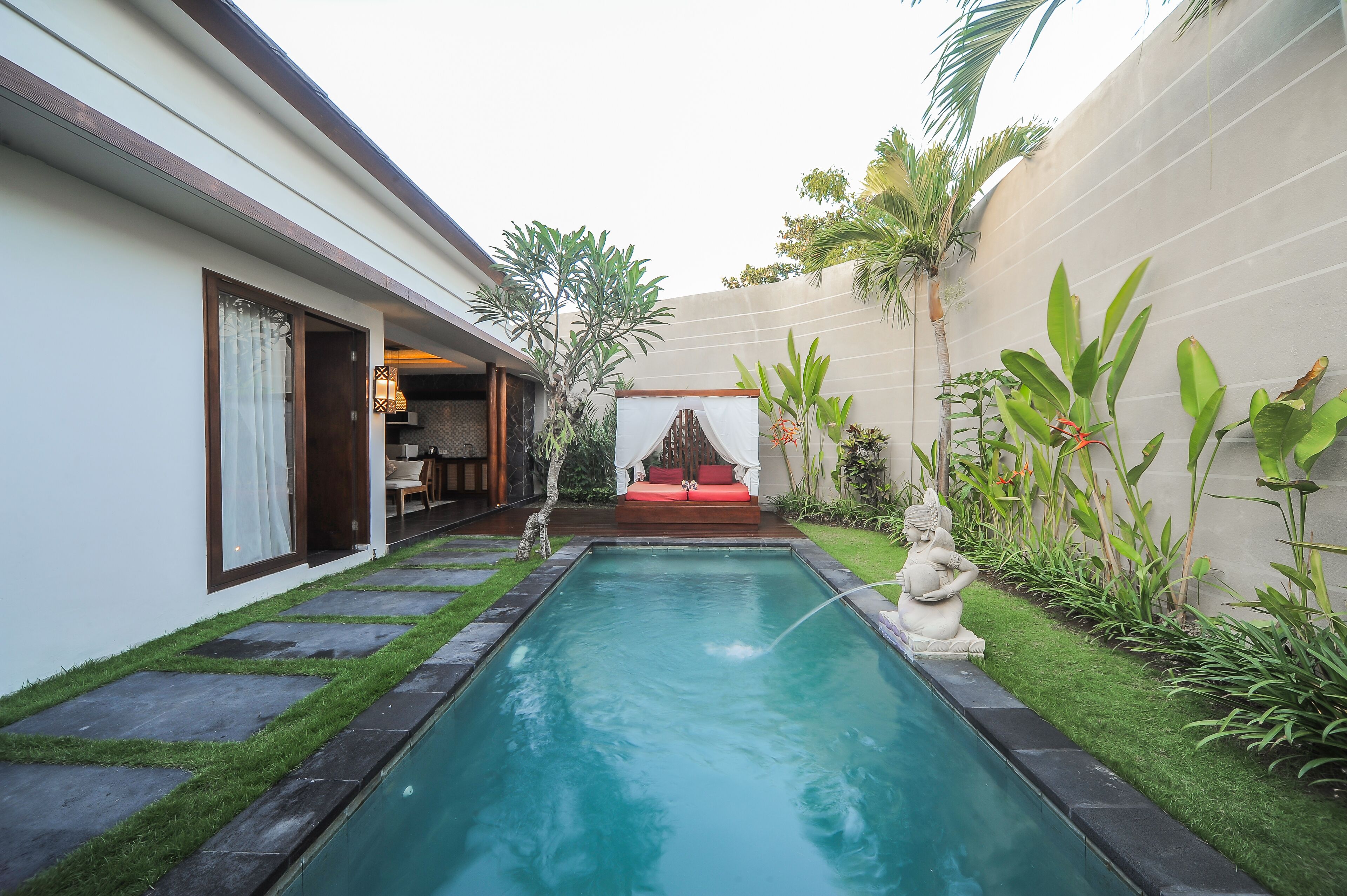Une piscine agrémentée d’une petite cascade, entourée d’un jardin bien entretenu où poussent différentes espèces de plantes, et comprenant un espace détente avec un coussin rouge.