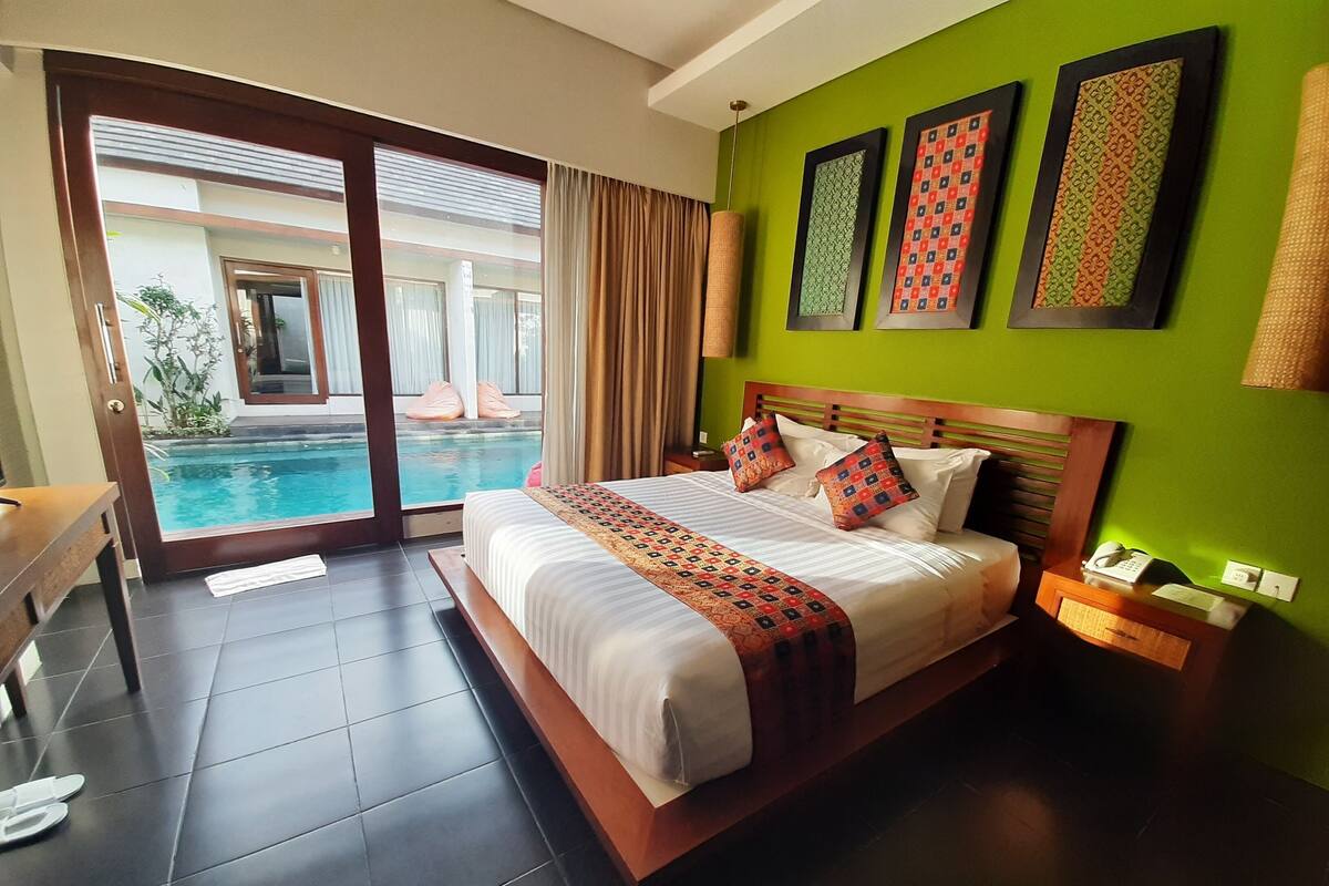 A hotel room with a large bed, a bedside table, a nightstand, a telephone, and a view of a swimming pool through a glass door.