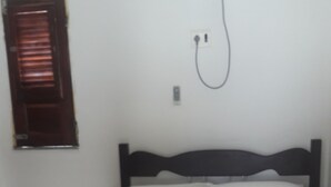 A bed with a beige bedspread, a wooden headboard, and white pillows. There is a power outlet on the wall and a window with blinds to the left.
