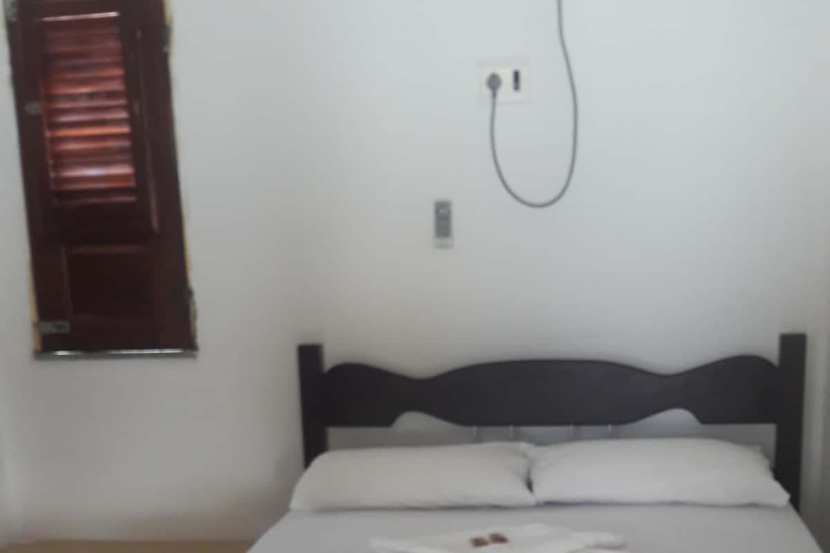 A bed with a beige bedspread, a wooden headboard, and white pillows. There is a power outlet on the wall and a window with blinds to the left.