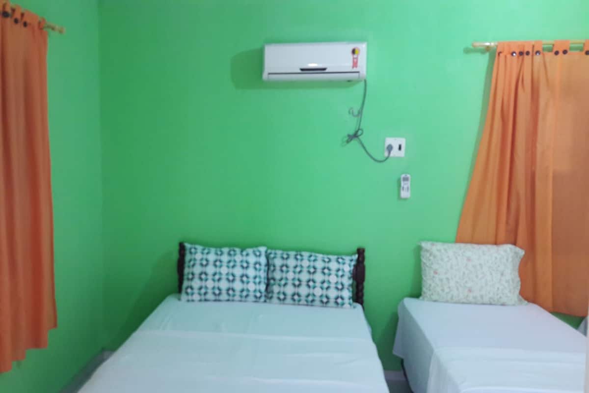 A room with two beds, one with patterned pillows and the other with plain pillows. There is an air conditioning unit on the wall.