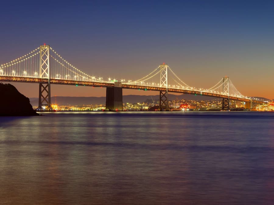 Vista nocturna de un puente colgante iluminado que cruza un cuerpo de agua, con las luces de la ciudad reflejándose en la superficie.