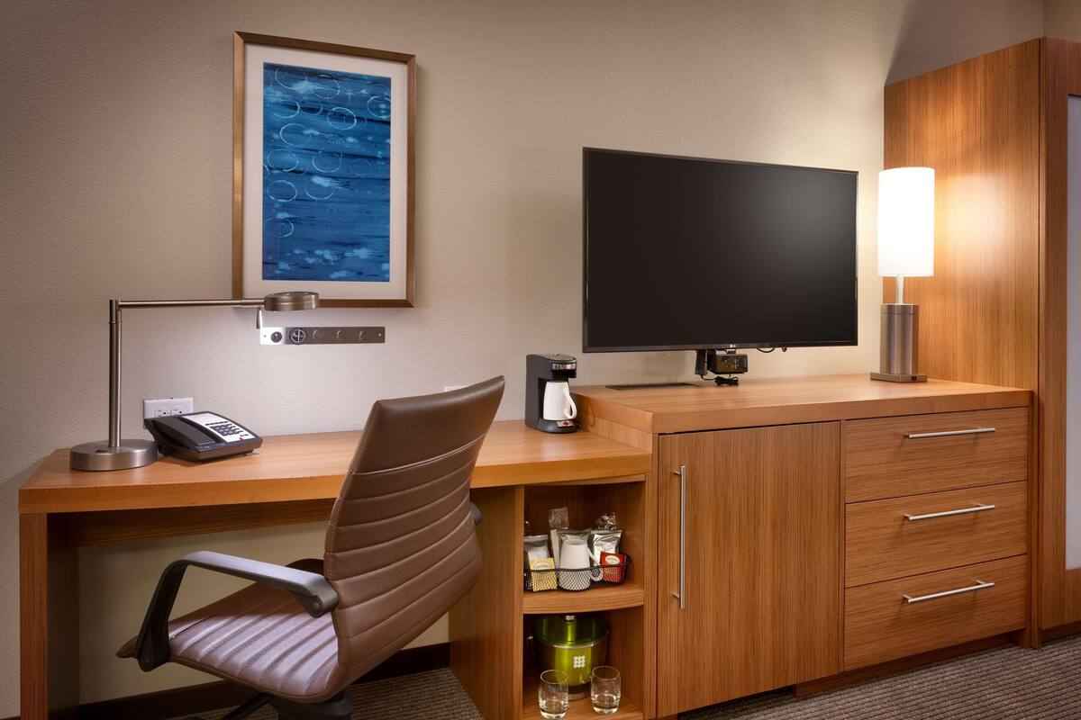 Habitación de hotel con un escritorio de madera, una silla, un televisor de pantalla plana, cafetera, lámpara y un mueble con cajones.
