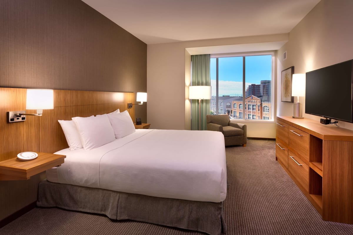 Habitación de hotel con una cama grande, lámparas de noche, un televisor, un escritorio con lámpara y vistas a la ciudad a través de la ventana.