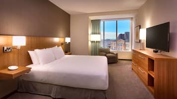 A hotel room with a large bed, bedside lamps, a TV, a desk with a lamp, and a view of the city through the window.