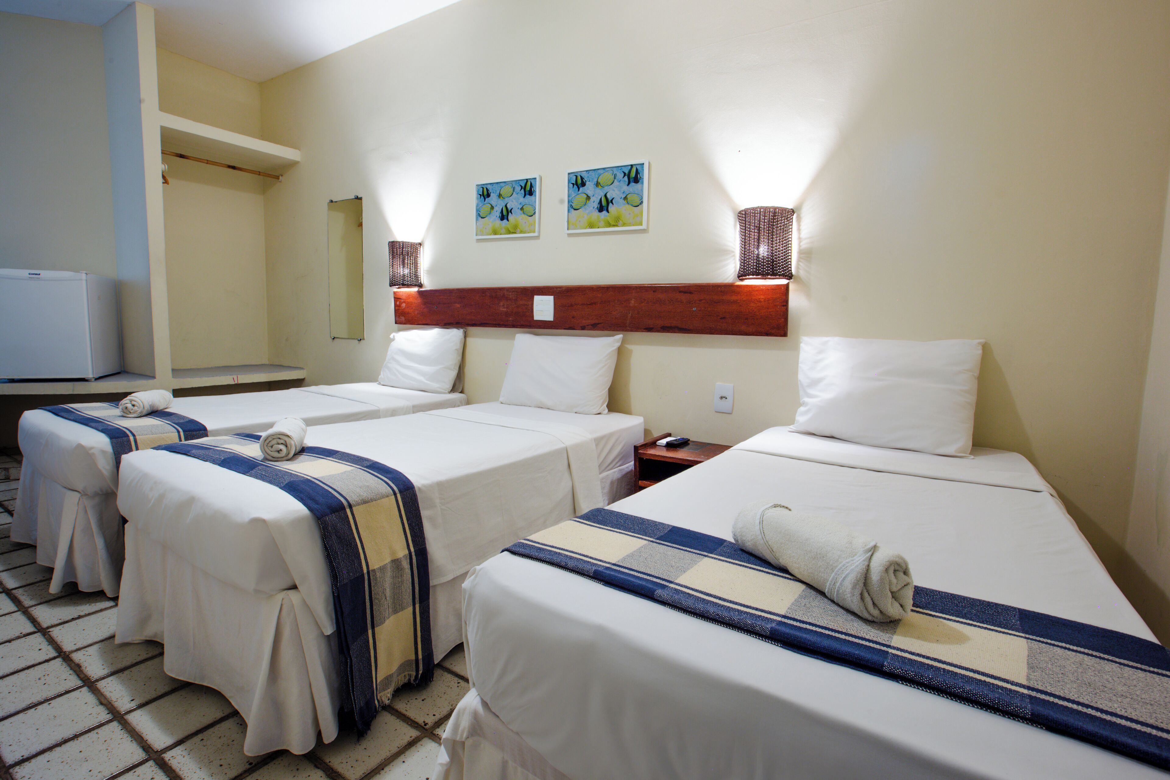 Quarto de hotel com três camas de solteiro, cabeceira de madeira, uma mesinha e uma televisão.