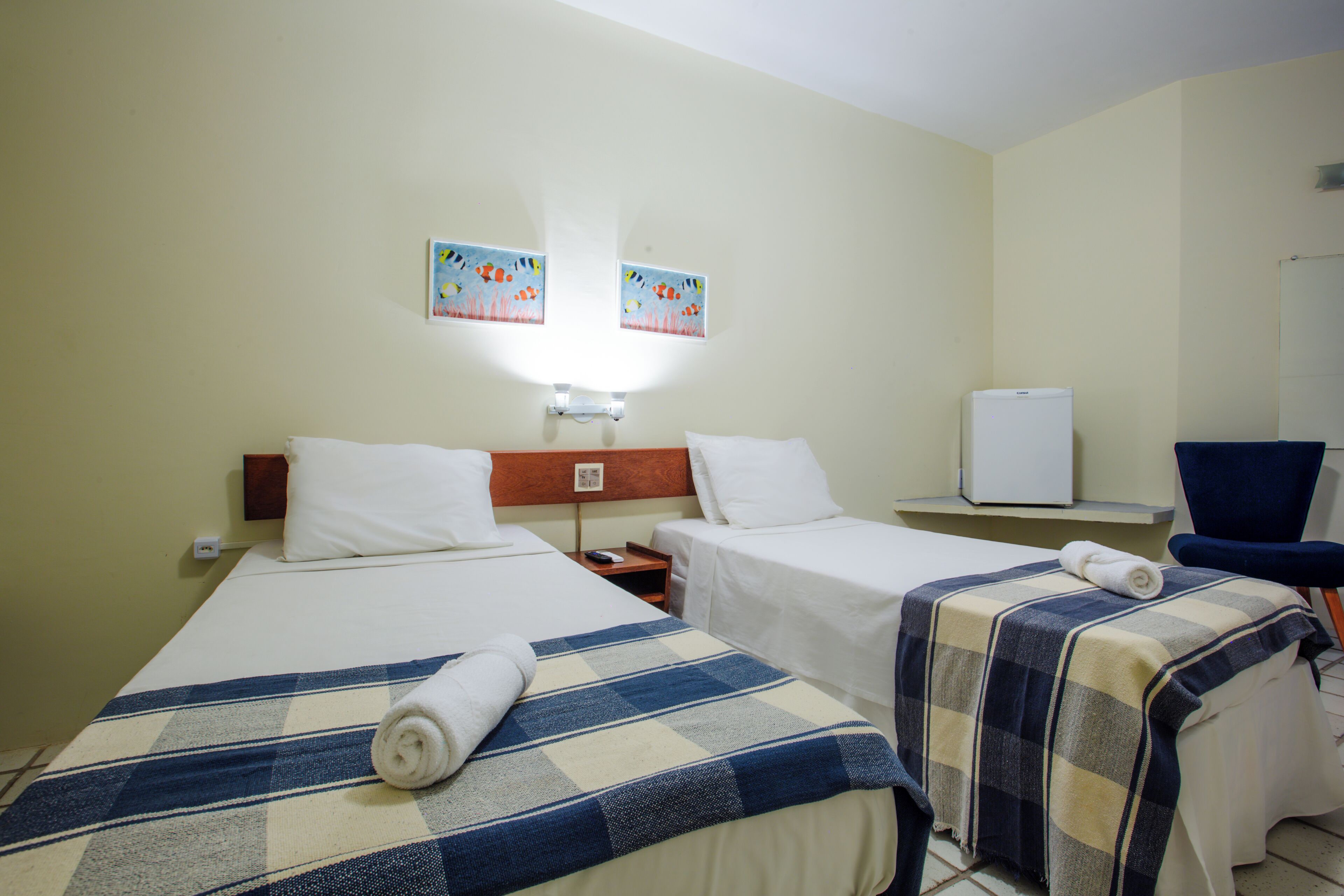 Habitación de hotel con dos camas individuales, una mesita pequeña, una silla, un refrigerador y dos cuadros colgados en la pared.