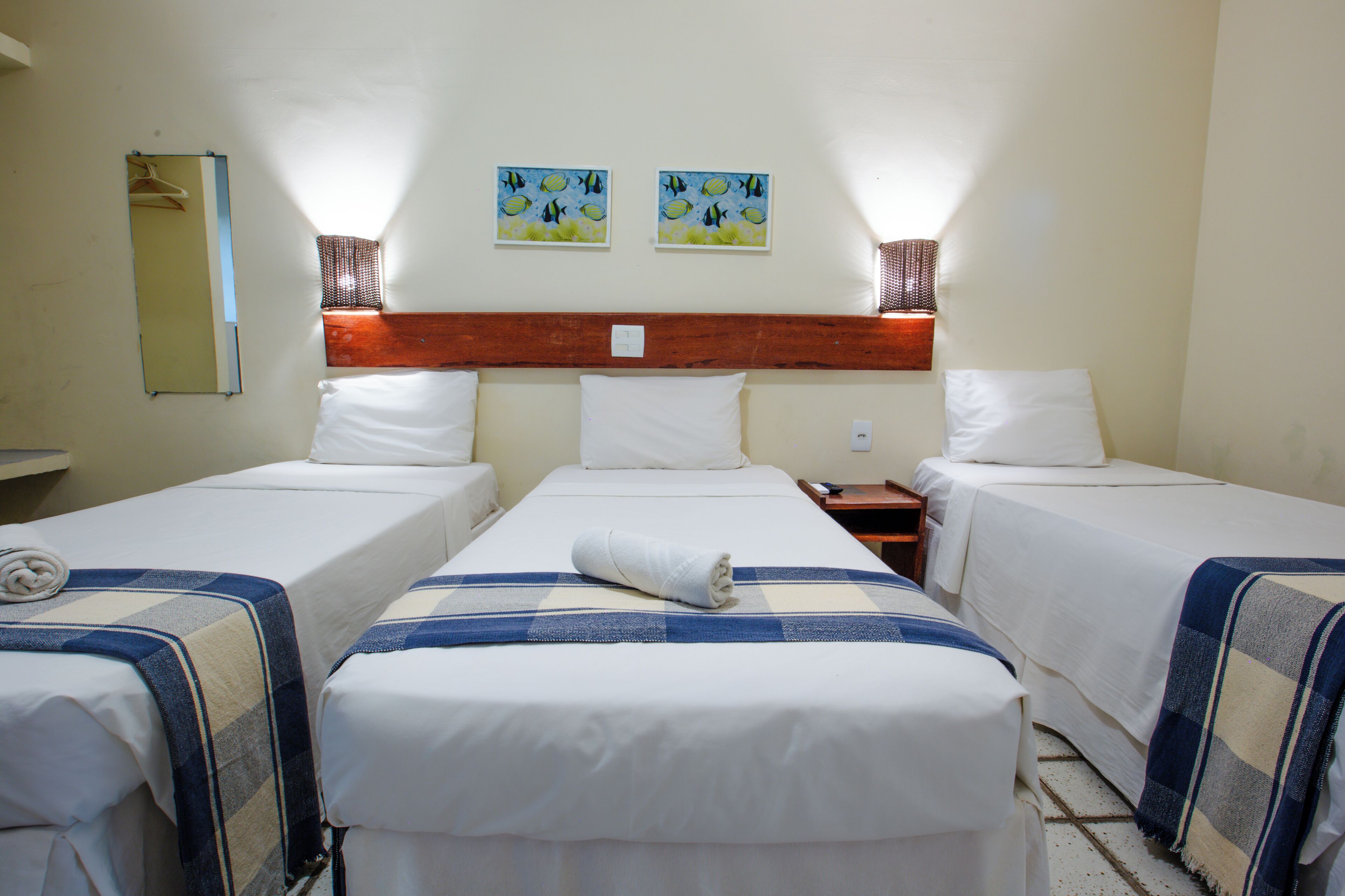 Une chambre d’hôtel avec trois lits simples, chacun recouvert d’une couverture à carreaux et agrémenté d’une serviette roulée. Deux tableaux sont accrochés au mur au-dessus des lits, ainsi qu’une tête de lit en bois et un miroir sur le mur de gauche.