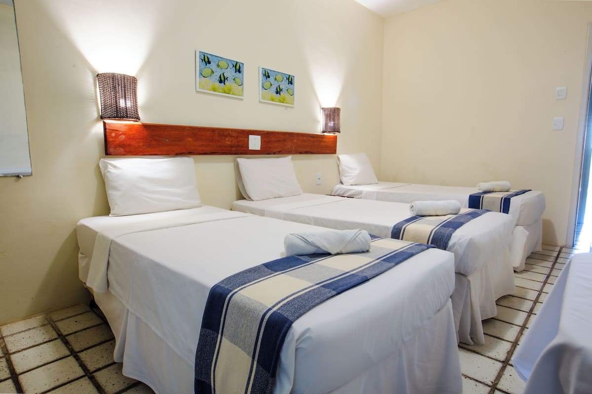 Ein Hotelzimmer mit vier Einzelbetten, jeweils mit einer weißen Matratze und einer blau-weiß gestreiften Decke. An beiden Seiten der Betten befinden sich jeweils zwei Wandlampen, darüber hängen zwei Bilder.