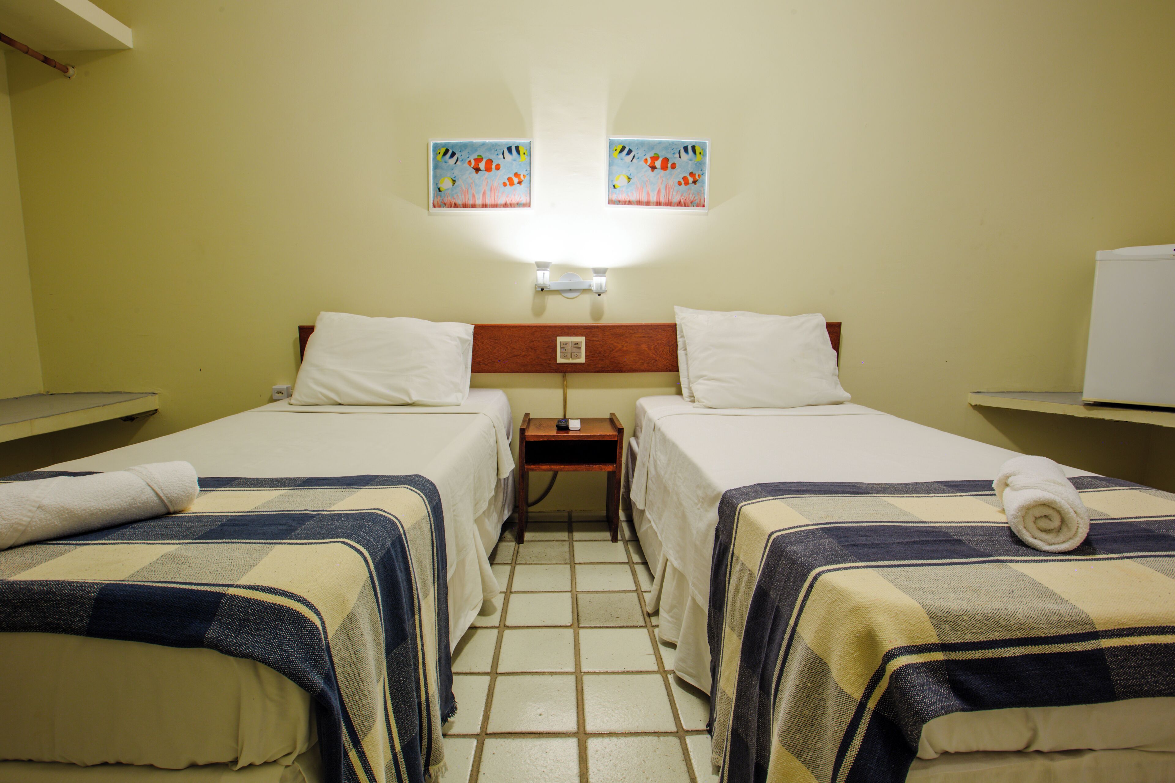 Ein Hotelzimmer mit zwei Betten, jedes mit einem weißen Kopfkissen und einem zusammengerollten Handtuch. Zwischen den Betten steht ein Nachttisch, und es gibt einen kleinen Tisch mit einer Lampe. Der Raum hat einen gefliesten Boden und helle Wände.
