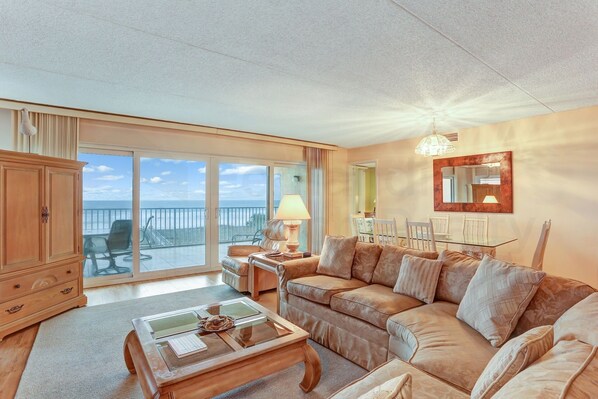 A spacious living room with a large sofa, a coffee table, and a dining area with a table and chairs. - Oceanfront Condo with Spacious Patio and Private Walkway to the Beach by RedAwning (Fernandina Beach)