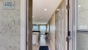 A hallway with a sign reading "102 Coral Sands," leading to a kitchen area with a dishwasher, a chair, and a window with curtains. - Coral Sands 102 2 Br condo by RedAwning (Fernandina Beach)