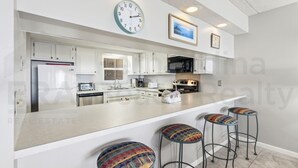 A modern kitchen with white cabinets, a central island with stools, a microwave, and a refrigerator. - Ocean View Condo, Easy Acces to the Pool and Private Walkway to the Beach by RedAwning (Fernandina Beach)
