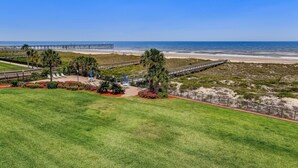A coastal view with a grassy area, palm trees, a wooden boardwalk, and a beach in the background. - Ocean View Condo, Easy Acces to the Pool and Private Walkway to the Beach by RedAwning (Fernandina Beach)