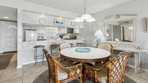 A modern kitchen with white cabinetry, a dining area with wicker chairs, and a large mirror above the counter. - Ocean View Condo, Easy Acces to the Pool and Private Walkway to the Beach by RedAwning (Fernandina Beach)
