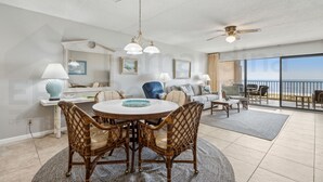 A spacious living room with a round dining table, wicker chairs, and a ceiling fan. There is a large window offering a view of the ocean. - Ocean View Condo, Easy Acces to the Pool and Private Walkway to the Beach by RedAwning (Fernandina Beach)