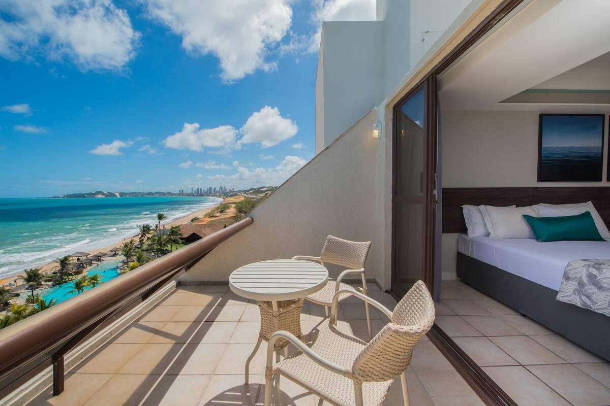 A balcony with a view of the beach and ocean, a bed with white bedding, and wicker furniture.